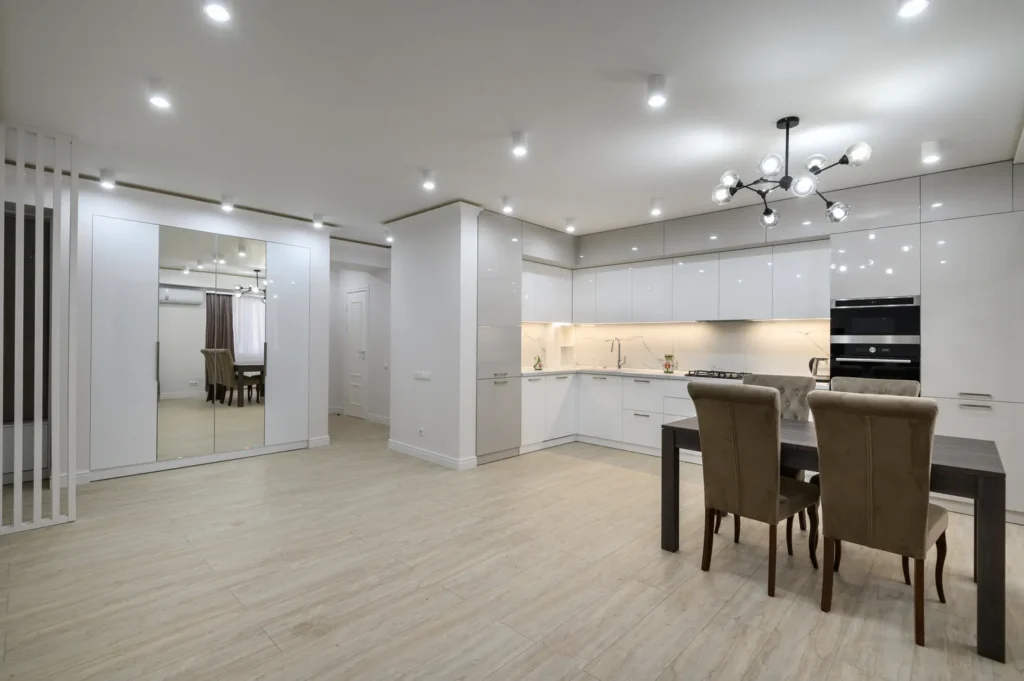 Cocina moderna y luminosa con muebles blancos y acabados elegantes, mesa de comedor de madera y sillas tapizadas en un ambiente minimalista.