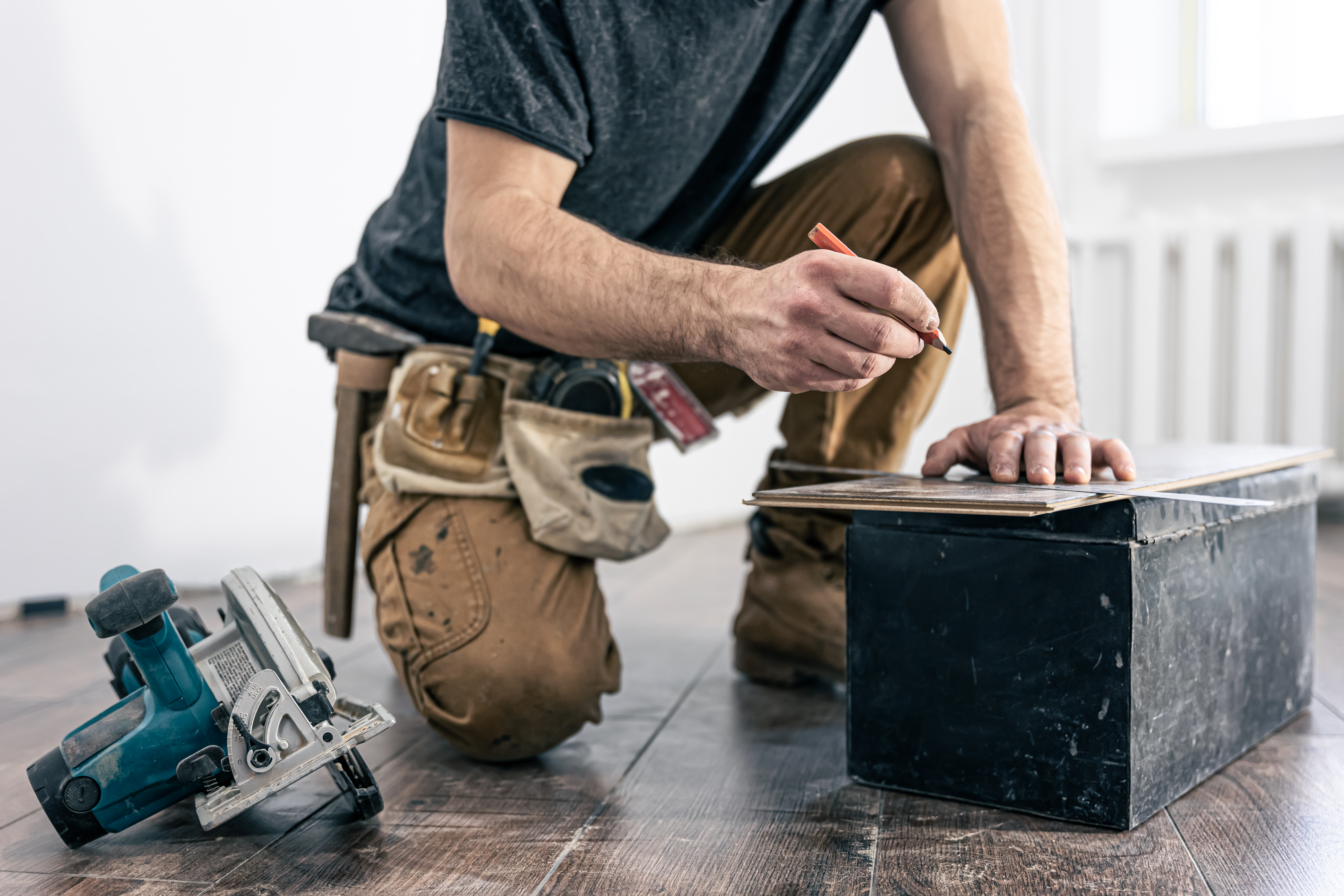 Hombre de obras de construcción midiendo y marcando una tabla de madera en un entorno de trabajo. En el fondo, se observa una sierra circular y un suelo de madera.