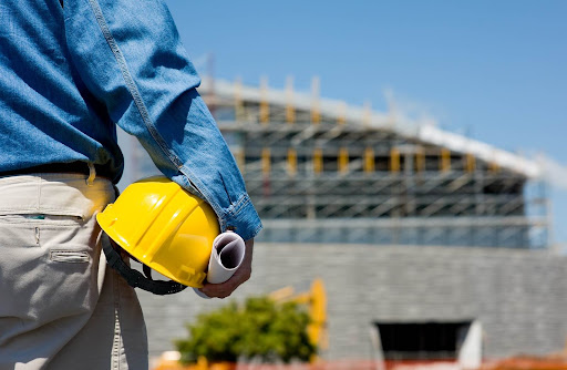 Hombre de pie con casco de seguridad amarillo y planos en la mano, observando una construcción en progreso.