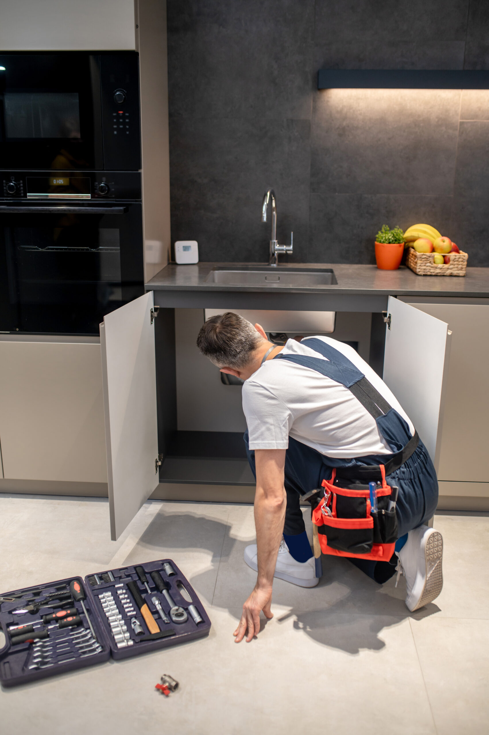 Técnico de mantenimiento revisando el interior de un mueble de cocina, con herramientas organizadas en una caja a su lado.