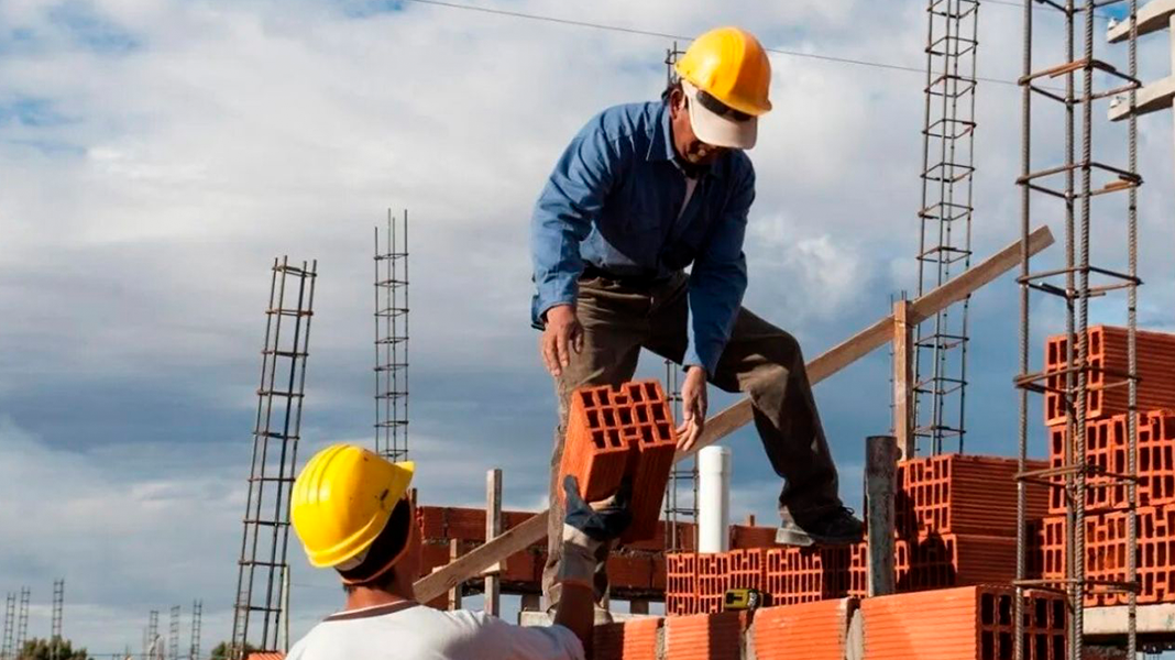 Trabajadores de la construcción en un sitio de obra, colaborando en el manejo de bloques de ladrillo, con estructuras de acero en el fondo y un cielo nublado.
